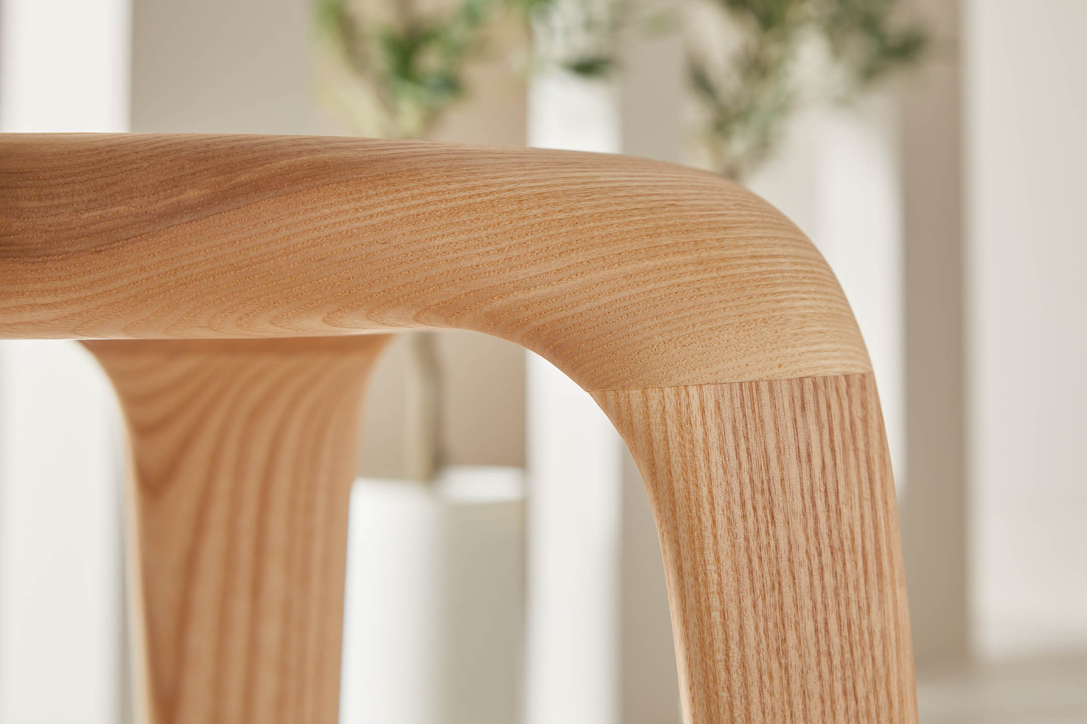 Close-up side profile of ash tripod stool seat, focused on the grain and joint where it meets the leg